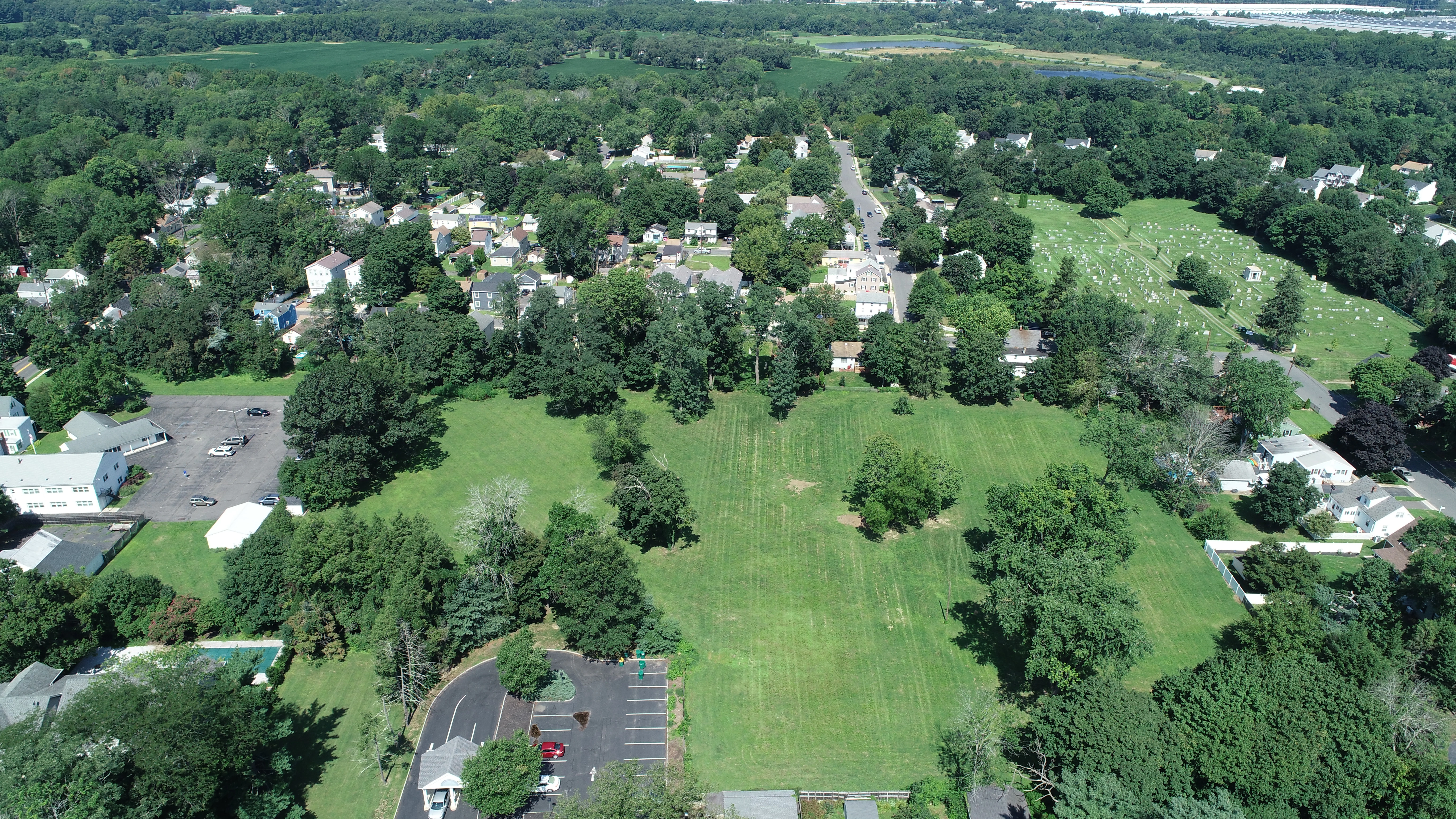 Aerial view of Memorial Park, where trees surround and scatter about neatly-trimmed grass. The park has an accessible parking lot at the front, and sits against a line of houses.